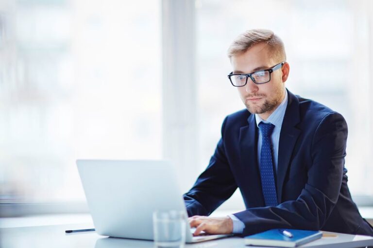 Man reviewing commercial roof inspection reports on laptop in office setting.
